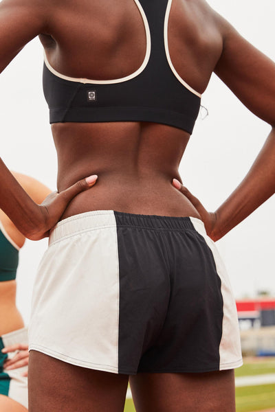 Female model wearing the Women's Rory Short in the Black contrast, standing in front of a track, shown from the back view.