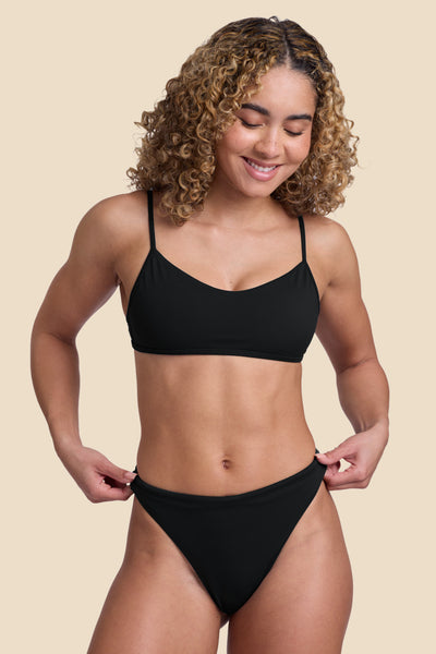 Female model wearing the Women’s Ava Bikini Bottom in the Black solid, standing in front of a yellow background, shown from front view. 
 