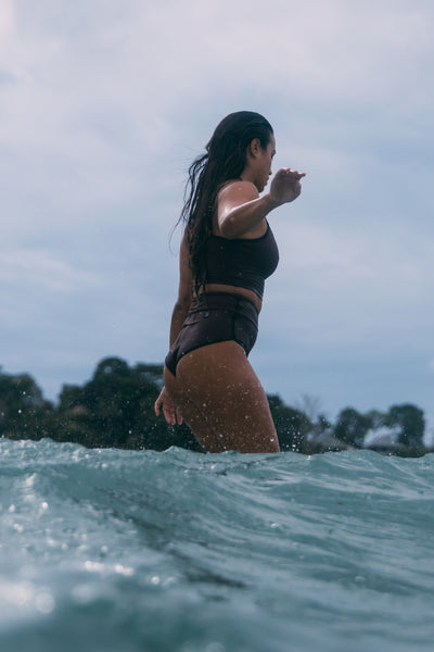 Female model wearing the Women's Aveline Bikini Bottom in the Sangria solid, standing on a surfboard in the ocean shown from the side view

|| Size S, Hips 35"  