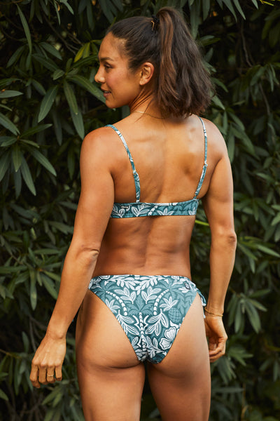 Female model wearing the Womens Bella Bikini Bottom in the Shorebreak print, standing in front of a green leaf background shown from the back view