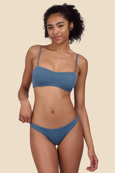 Female Model wearing the Women's Kelia Bikini Bottom in the Dolphins solid, standing in front of a yellow background, shown from the front view