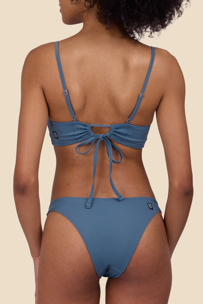 Female Model wearing the Women's Kelia Bikini Bottom in the Dolphins solid, standing in front of a yellow background, shown from the back view