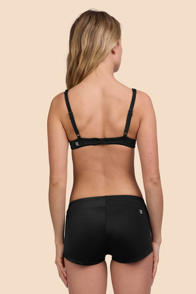 Female model wearing the Women's Piha Surf Short in the Black solid, standing in front of a yellow background shown from the back view

|| Size S, Hips 36"
