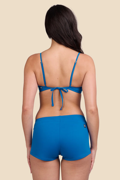 Female model wearing the Women's Piha Surf Short in the Blue Moon solid, standing in front of a yellow background shown from the back view

|| Size S, Hips 34"  
