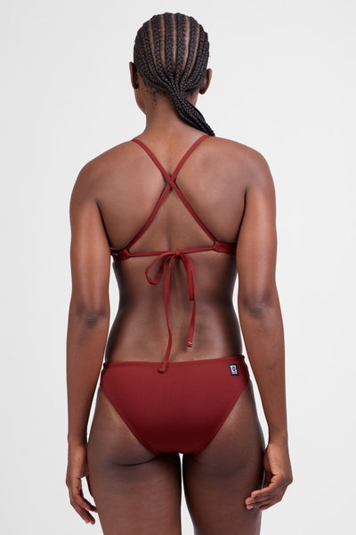 Female model wearing the Women’s Andy Bikini Bottom in the Paprika solid, standing in front of a grey background, shown from back view. 
