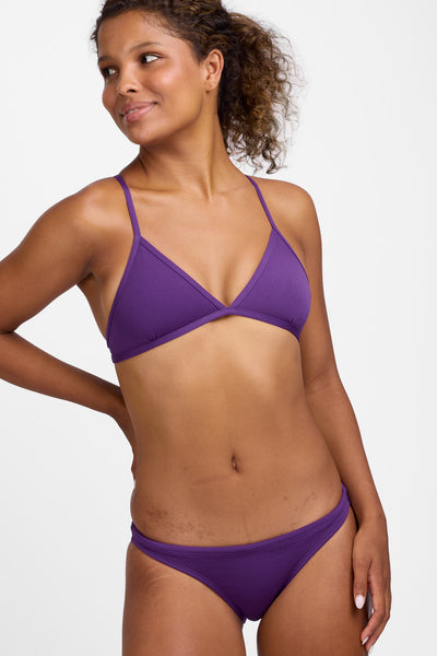 Female model wearing the Women’s Europe Bikini Bottoms in the Grape solid, standing in front of a grey background, shown from front view. 
