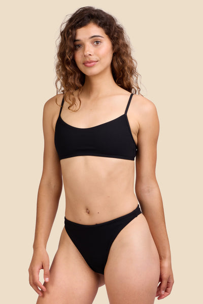 Female model wearing the Women's Ava Bikini Bottom in the Black Ribbed solid, standing in front of a yellow background shown from the front view

|| Size S, Hips 39"