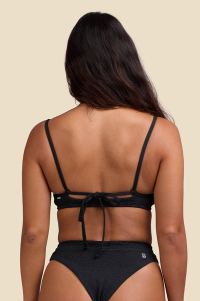 Female model wearing the Women's Delphine Bikini Top in the Black solid, standing in front of a yellow background shown from the back view

|| Size S, Bust 34"  