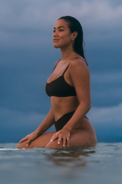 Female model wearing the Women's Delphine Bikini Top in the Sangria solid, sitting on a surfboard in the ocean shown from the side view

|| Size S, Bust 34"  
