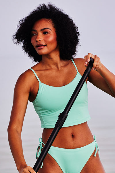 Female model wearing the Women's Luca Bikini Top in the Mint Tulip solid, standing in top of a paddle board in front of the ocean shown from the front view