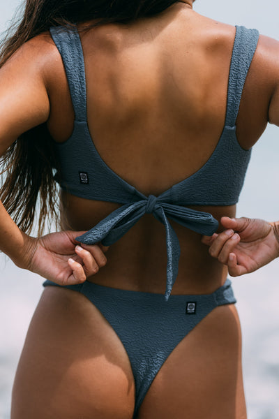 Female model wearing the Women's Lyra Bikini Top in the Horizon solid, standing in front of a beach shown from the back view

|| Size S, Bust 34"  