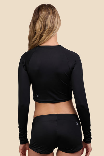 Female model wearing the Women's Moana Long Sleeve Crop Surf Rashie in the Black solid, standing in front of a yellow background shown from the back view

|| Size S, Bust 33"  