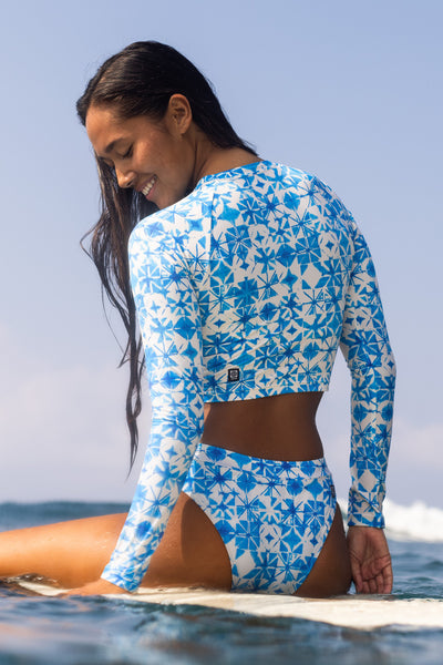 Female model wearing the Women's Moana Rashie in the Bluefin print, sitting on a surfboard with ocean in the background shown from the back view