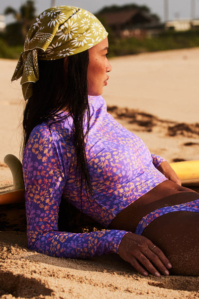 Female model wearing the Women's Moana Long Sleeve Crop Rashguard in the Jellybean print, laying in front of a beach shown from the side view