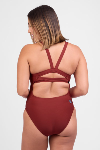 Female model wearing the Women’s Caroline One Piece in the Paprika solid, standing in front of a grey background, shown from back view. 
