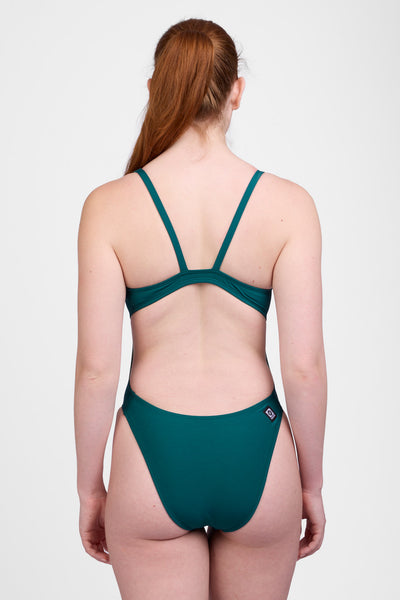 
Female model wearing the Women’s Christie One Piece Swimsuit in the Peacock solid, standing in front of a grey background, shown from back view. 
