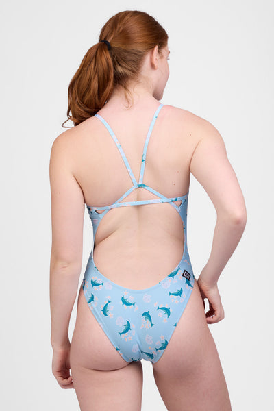 Female model wearing the Women’s Perry One Piece Swimsuit in the Dolphrenz print, standing in front of a grey background, shown from back view. 