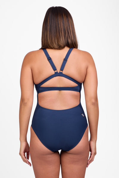 
Female model wearing the Women’s Rylee One Piece Swimsuit in the Navy solid, standing in front of a grey background, shown from back view. 
