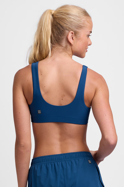 Female model wearing the Women's Arabella Sports Bra in the Blue Flame solid, standing in front of a grey background, shown from the back view.