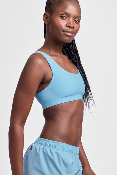 Female model wearing the Women's Arabella Sports Bra in the Sky Wave solid, standing in front of a grey background, shown from the side view.