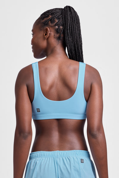 Female model wearing the Women's Arabella Sports Bra in the Sky Wave solid, standing in front of a grey background, shown from the back view.