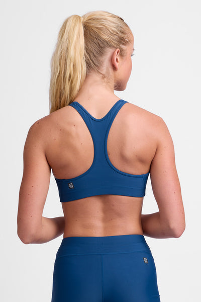 Female model wearing the Women's Molly Sports Bra in the Blue Flame solid, standing in front of a grey background, shown from the back view.